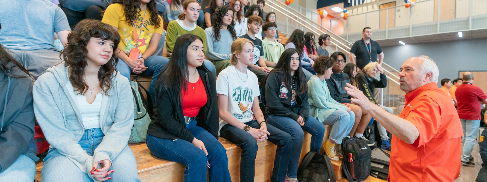 President Bailey speaks in front of a large group of students seated in rows of benches, their attentive faces turned towards him.